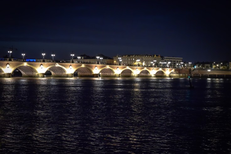 An illuminated bridge with many arches spans the river in Bordeaux at night