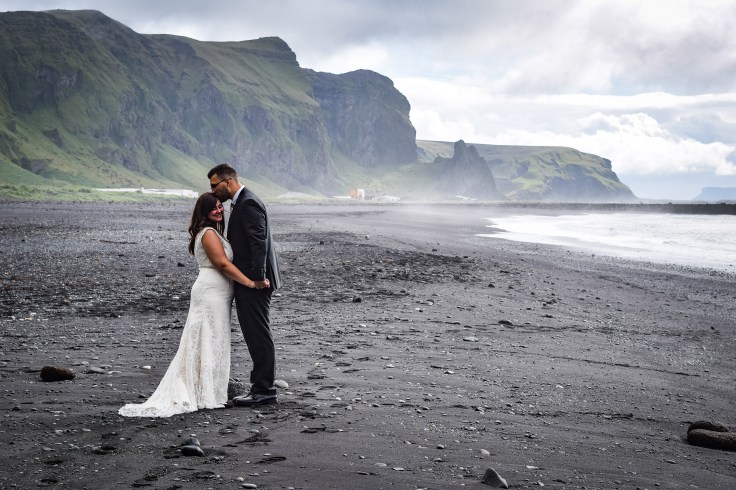 My brother-in-law kissing my sister on the forehead in front of the mountains on a black sand beach